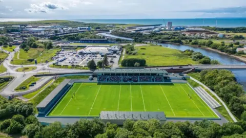 Cumberland Council Artist's impression showing Cumberland Sports Village from above. It has stands on three sides, each with a roof, and a terrace area on the fourth. The pitch is laid out for a rugby match with tall posts at either end. The River Derwent runs along the right-hand side of the ground. In the background are Workington's Tesco supermarket and roads leading into the town centre.