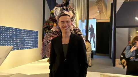 Charles Jeffrey, a young smart-dressed man in a black jacket smiles in front of the exhibition at the V&A.