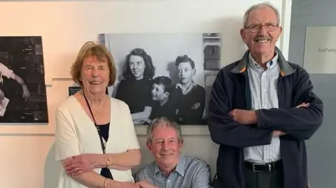 Steven Howells A woman and two men, who are smiling at the camera, are standing in front of a black and white photograph of themselves taken decades earlier by Bob Howells.