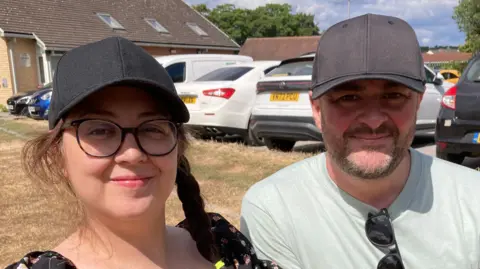 BBC Bryony Hill and James Hill, a couple in their 40s, wearing black caps on a sunny day in Nottinghamshire. They are both smiling as they sit on a community playing field. A community centre and car park can be seen behind the couple. 