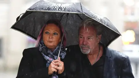 PA Media Harry Dunn's mother and stepfather dressed in black and both huddling under an umbrella on a rainy day