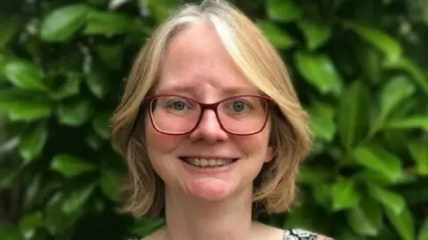Mai Black Mai Black smiles at the camera as she stands in front of a large green bush. She has short blonde hair and red glasses.