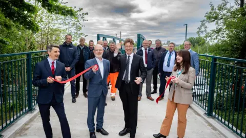 Leeds City Council Four smartly dressed people hold red ribbons and smile while stood on a footbridge. A larger group of people, almost all of them men, standing behind them in the background.