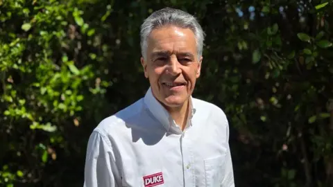 Duke Marketing Peter Duke has short grey hair, he wears a white shirt with a red Duke logo embroidered on his right chest. He stands in front of some green bushes.