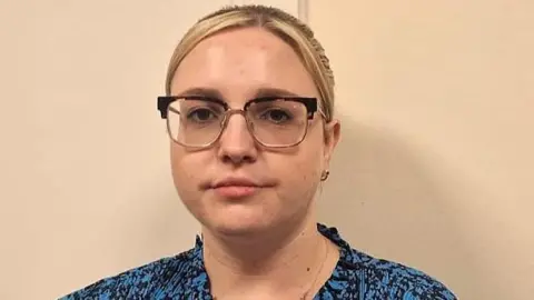 Sophie has blonde hair and wears glasses. She is wearing a blue blouse and looking at the camera. 