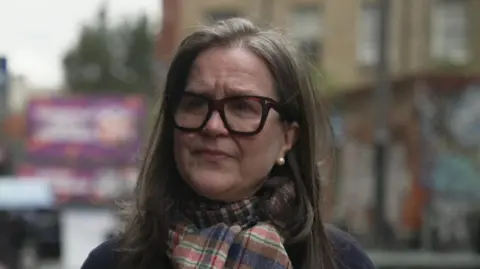 Jenny Black looking past the camera. She has long hair and is wearing a chequered scarf and glasses with a dark frame.