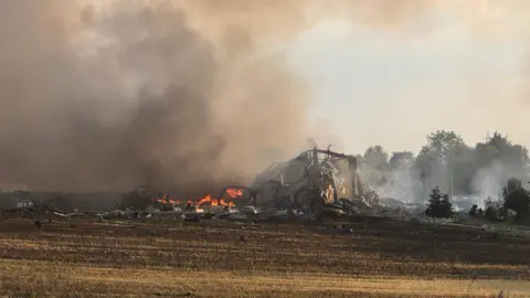 Zoie O'Brien/BBC A collapsed building surrounded by fire and smoke on a field.