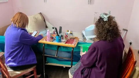 Martin Giles/BBC Clare Reynolds is sitting at a bed in a room with pink walls. She is wearing a purple fleece and reading her phone to a woman in the bed. Next to Clare Reynolds is Rhiannon Reed who is wearing a dark purple cardigan and blue spotted dress. She is filing away paperwork. The face of the woman in the bed has been blurred out. 