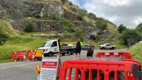 A winding road in Cheddar Gorge which has been closed with plastic fencing and orange cones. There is an A-board sign on the side of the road which says Caution, filming in progress. There are two men stood in the middle of the road, one with his arms crossed and the other wearing a hi-vis vest.