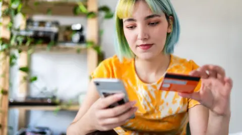 Getty Images A young woman with blue, green and yellow hair - wearing an orange, tie dyed t-shirt - holds her smartphone in one hand and a credit card in the other. A bookshelf covered in plants and ornaments is in the blurred background behind her.