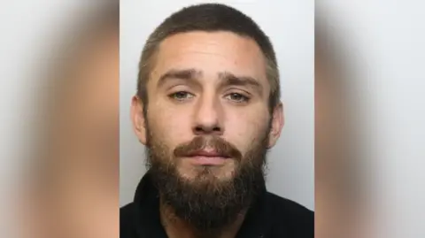 Custody image of Rikki Holmes. He looks forward at the camera, wearing a black T-shirt and facial hair