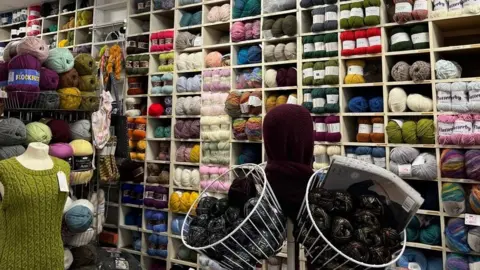 Bundles of yarn in all colours fill shelves in the shop. There are hanging baskets with yarn and a mannequin wearing a green jumper.