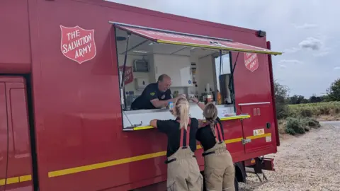 Salvation Army A large red canteen trailer with The Salvation Army branding, a man inside is handing cups to two firefighters.