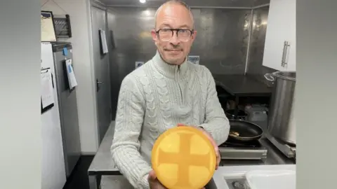 Matthew Lloyd inside his cheese factory. He is wearing a beige jumper with black glasses, and is holding a large, yellow wheel of cheese