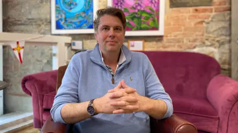 A man with dark brown hair, wearing a light blue jumper sits in an arm chair.