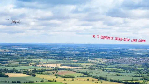 A small plane is in the sky over some countryside, behind it is a banner which reads 'No to Corporate Greed - Stop Lime Down.'