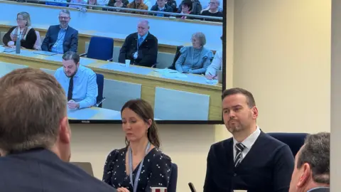 BBC/Simon Dedman A council meeting with a man who is the council leader sat to the right of the frame listening. He has a short haircut and trimmed beard. A female council officer sat next time is in the centre of the picture. Behind them is a TV screen showing the live feed of the meeting which has a councillor in the centre speaking from notes.