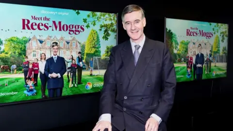 PA Media Jacob, smiling, wearing a blue jacket and tie, sitting in front of some posters for the TV show Meet the Rees-Moggs