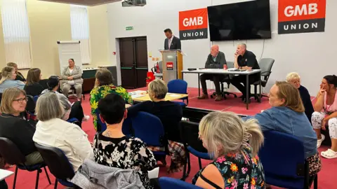 A man talks at a podium set up on a stage in a meeting room. People are sitting down on chairs in the foreground watching him speak.
