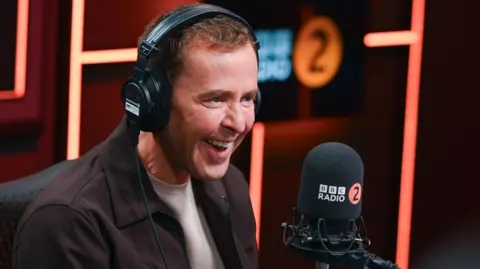 Presenter Scott Mills dengan headphone di depan mikrofon di BBC Radio 2 Breakfast Show Studio