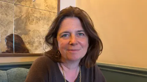 A person sits at a table in a softly lit café or restaurant, facing the camera. The person is wearing a brown top and a gold chain necklace. Behind the person is a wall with a decorative mirror that reflects part of the room.