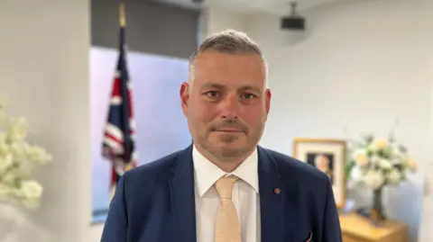 A close-up portrait of a man with short grey hair and a beard, wearing a navy blue suit jacket, white shirt, and gold-patterned tie, standing in an office setting with a Union Jack flag blurred in the background.