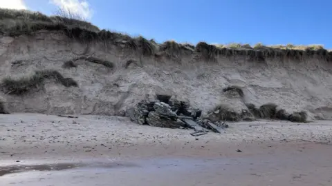BBC / Luke Walton A long sand dune, topped with grass, borders a sandy beach in the foreground. A grey concrete structure is poking out of the dune. Sand bags filled with concrete create the walls of a hut-shaped structure. There are concrete slabs that appear to have collapsed in front of it.