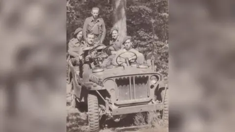 Family handout Jim Almond is sitting behind a Vickers machine gun mounting in the front of a Land Rover. Four other SAS men are in the vehicle. They are pictured in a clearing in a forest, with dense woodland in the background.