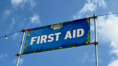 A first aid banner at Kendal Calling. It is blue, with white lettering and has a Kendal Calling logo. It is set high on poles, against the blue sky. 