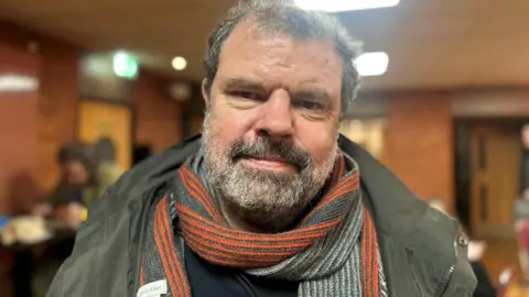 Adam Lee smiling at the camera, has dark grey hair and a beard, is wearing a green coat with a grey and orange scarf.