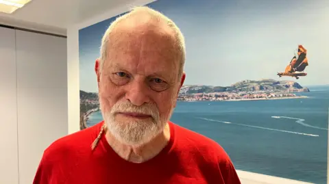 Terry Gilliam smiles at the camera. He has blue eyes and a silver beard. He wears a red t-shirt. Behind him, an image of a coastline can be seen on a wall. 