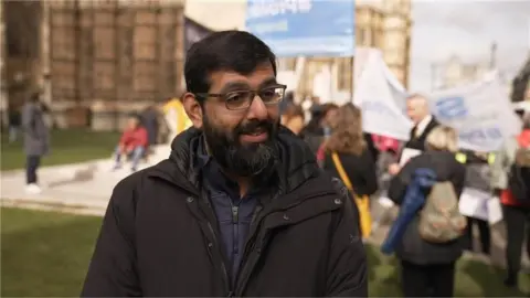 An Asian man with a beard and glasses stands in front of demonstrators outside parliament holding placards. He wears a black coat.