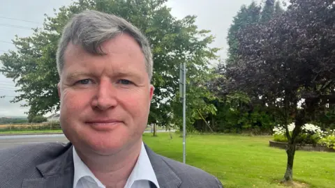 Malcolm Byrne Man with grey hair wearing a white shirt and grey jacket. He is pictured in front of a green area and fields.