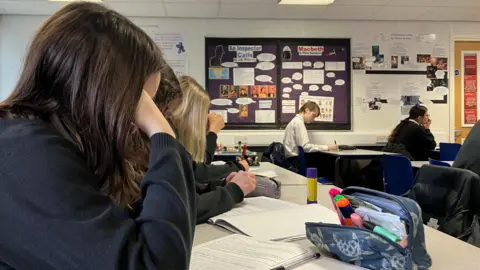 There are a row of students in a classroom in navy jumpers with their heads down looking at their notebooks. A pencil case is on the table. 