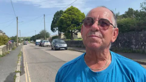 A man wearing a bright blue tshirt and sunglasses looks up at the cameraman while being interviewed about the road closure 