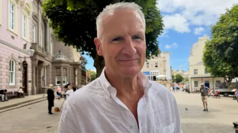 Ammar Ebrahim/BBC Senator Tom Binet is wearing a white shirt and standing outside in a sunny square. a man in shorts in in the background and a television crew are also interviewing someone dressed in black behind Senator Binet.