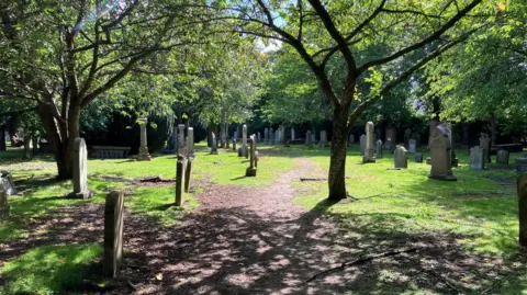 Gravestone under trees in Inverness' Chapel Yard Cemetery. The leaves of the trees cast dabbled shadows on the ground.