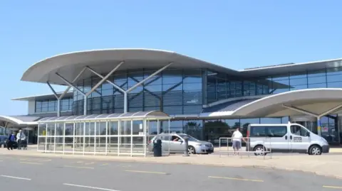 Guernsey airport main entrance. It is a glass building with vehicles parked out the front of it.