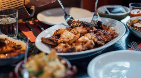 A person is using cutlery to lift meat out of a dish on a table, with other dishes, plates and glasses nearby.