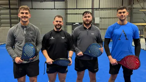 Becki Bowden / BBC Four man wearing sports shirts and shorts smile as they stand on a court holding padel raquets - plastic short-handled, solid raquets with holes through the surface. They are standing on a blue court in a warehouse-like building.