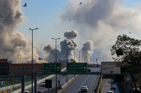 AFP via Getty Images Motorists drive along an expressway as plumes of smoke rise after a strike in Tehran on 5 March 2026.