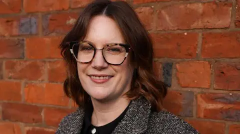 Black Country Living Museum A woman with shoulder-length brown hair and tortoiseshell glasses smiles at the camera. She is wearing a grey coat over a black top with gold buttons. She is standing in front of a red brick wall.
