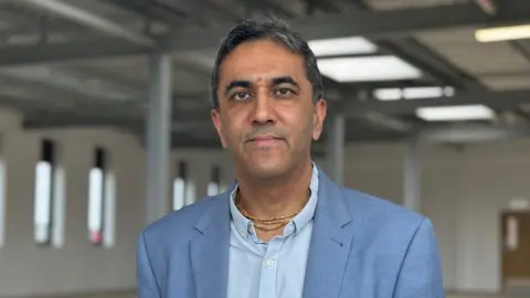 Dr Bhimpuria is wearing a blue suit and pale blue shirt. He is standing in a large open space with many windows and several pillars. This room will be converted and divided up to become a new GP surgery.