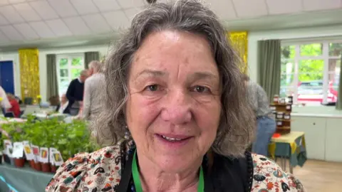 Tom Jackson/BBC Christa Jostock has shoulder length grey hair and is wearing a floral patterned T-shirt and a green lanyard. She is standing in a room which has plants and other items on display.