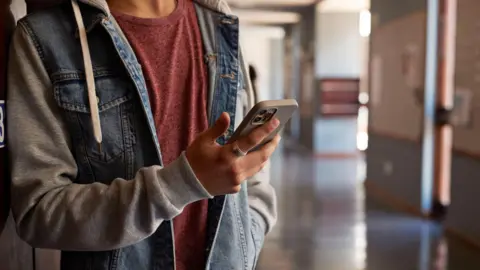 A close-up of an anonymous young person looking at a phone in a school corridor. They have a denim blue and grey jacket and a red T-shirt underneath, with a ring on the finger.