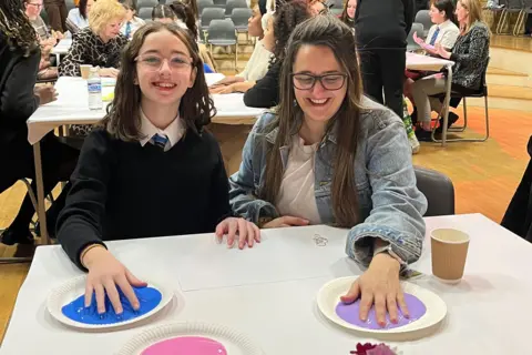 Discovery Educational Trust A girl in a black jumper, white shirt and tie is smiling at the camera, while sitting at a table. She has her hand on a paper plate, which has blue paint on it. To her left, is a woman in a denim blue shirt, who also has her hand on a paper plate with purple paint on it. She is smiling, looking down at the plate.