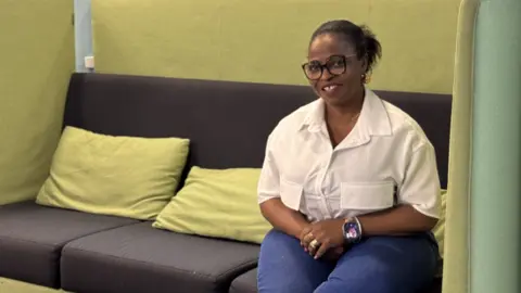 Christianah Abayomi-Daniel is sat on a large, brown and mustard-coloured settee. She is on the right of the settee and is wearing a white shirt and blue jeans. She has glasses on and her hair tied back.