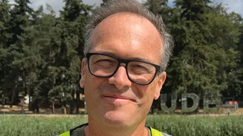 A head shot of a man wearing glasses. He is standing in a field with a mirrored Latitude sign behind him in the distance.