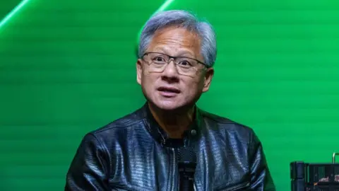 Getty Images Jensen Huang in signature black leather jacket in front of a green background holding a microphone and speaking questions at an event.