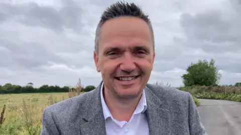 Mike Bell is standing on a rural road next to a field. He is wearing a grey suit jacket and a white shirt. He is smiling at the camera.